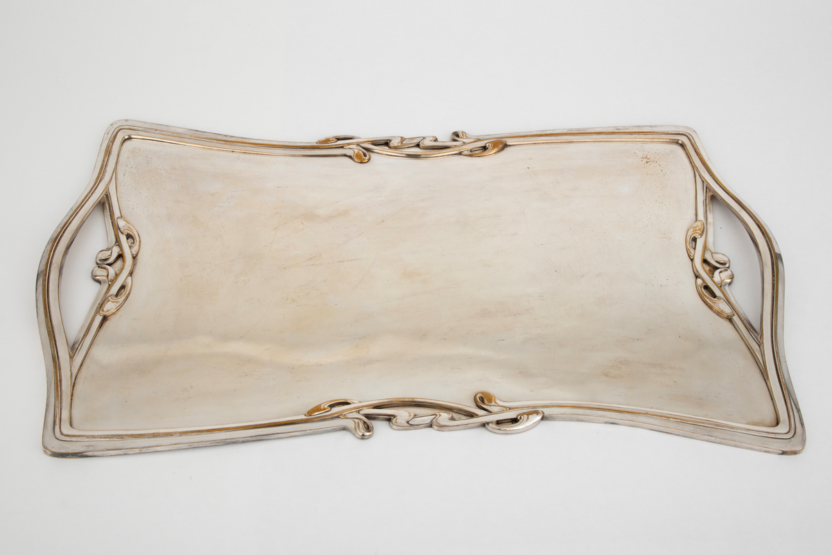 Flatt brett i sølv. Speilet er flatt, mens kantene er dekorert med art nouveau-profiler og ornamentikk. På hver kortside er det håndtak som er innfelt i kantens profil og ornamentikk.