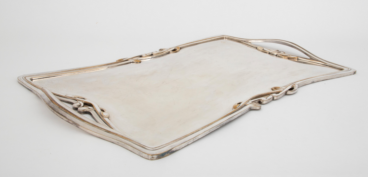 Flatt brett i sølv. Speilet er flatt, mens kantene er dekorert med art nouveau-profiler og ornamentikk. På hver kortside er det håndtak som er innfelt i kantens profil og ornamentikk.