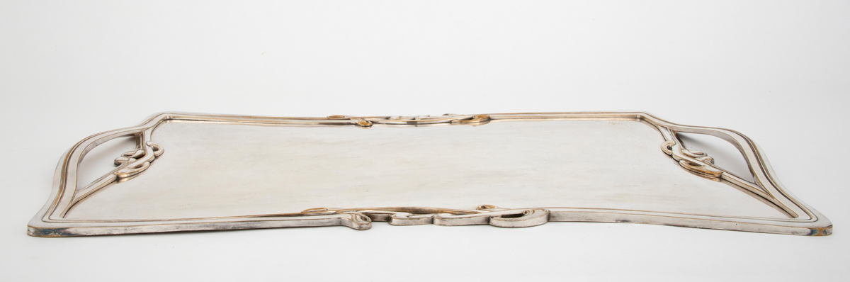 Flatt brett i sølv. Speilet er flatt, mens kantene er dekorert med art nouveau-profiler og ornamentikk. På hver kortside er det håndtak som er innfelt i kantens profil og ornamentikk.