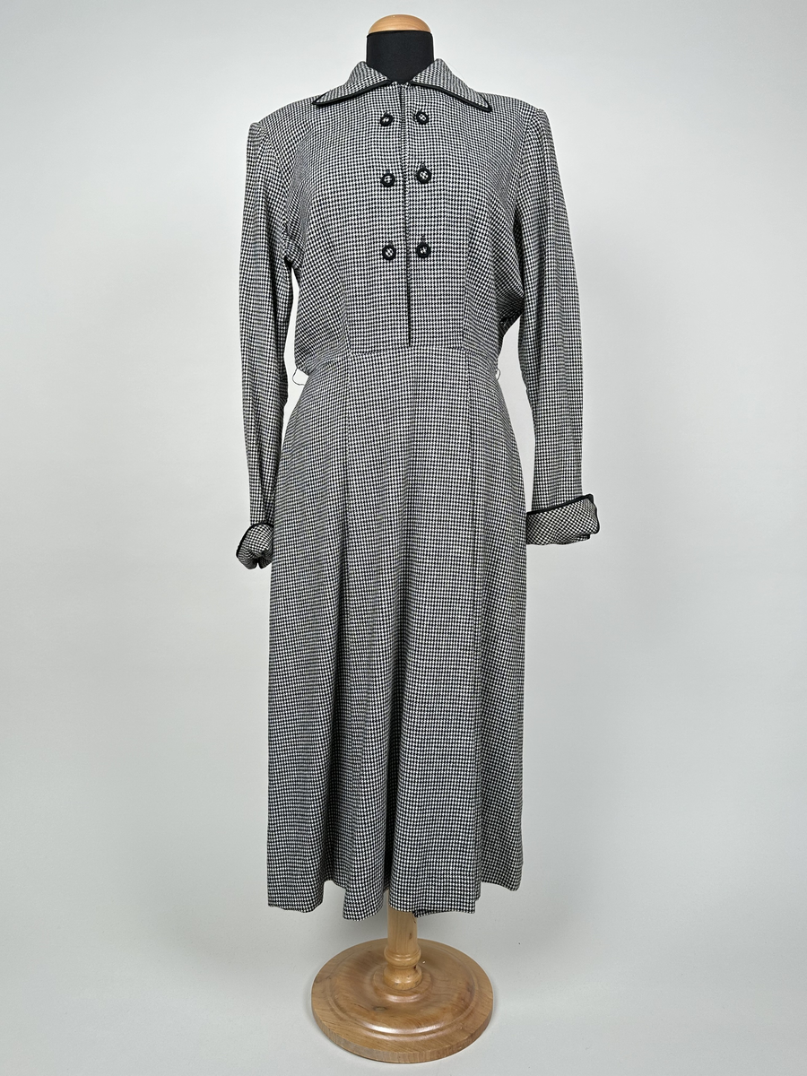 Klänning av hundtandsmönstrat ylletyg i svart och vitt. Krage och uppvikta manschetter kantade med svart band. Axelvaddar. Midjeskuren, med överdel som är öppen ner till midjan och försedd med isättning som är fäst i midjesömmen. Isättningen med dubbla knapprader och en tryckknapp överst, som knäppes till överdelen för att sluta öppningen. Tunna hällor för skärp på var sida om midjan. Dragkedja i sidan, under armhålan.