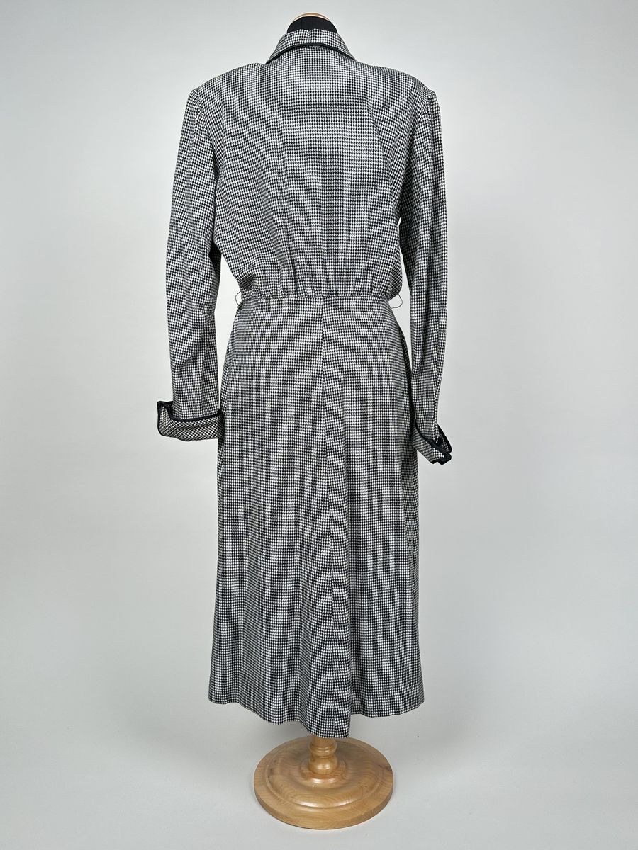 Klänning av hundtandsmönstrat ylletyg i svart och vitt. Krage och uppvikta manschetter kantade med svart band. Axelvaddar. Midjeskuren, med överdel som är öppen ner till midjan och försedd med isättning som är fäst i midjesömmen. Isättningen med dubbla knapprader och en tryckknapp överst, som knäppes till överdelen för att sluta öppningen. Tunna hällor för skärp på var sida om midjan. Dragkedja i sidan, under armhålan.