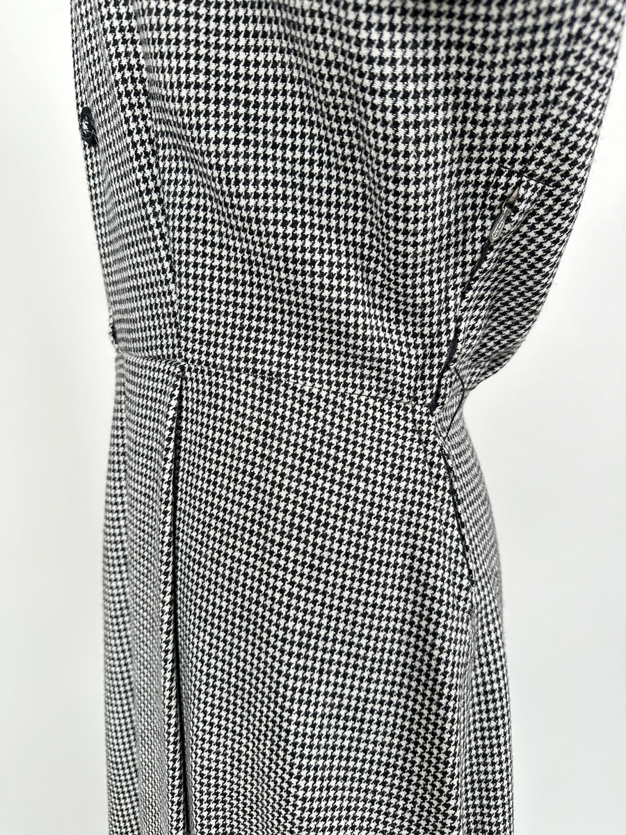 Klänning av hundtandsmönstrat ylletyg i svart och vitt. Krage och uppvikta manschetter kantade med svart band. Axelvaddar. Midjeskuren, med överdel som är öppen ner till midjan och försedd med isättning som är fäst i midjesömmen. Isättningen med dubbla knapprader och en tryckknapp överst, som knäppes till överdelen för att sluta öppningen. Tunna hällor för skärp på var sida om midjan. Dragkedja i sidan, under armhålan.