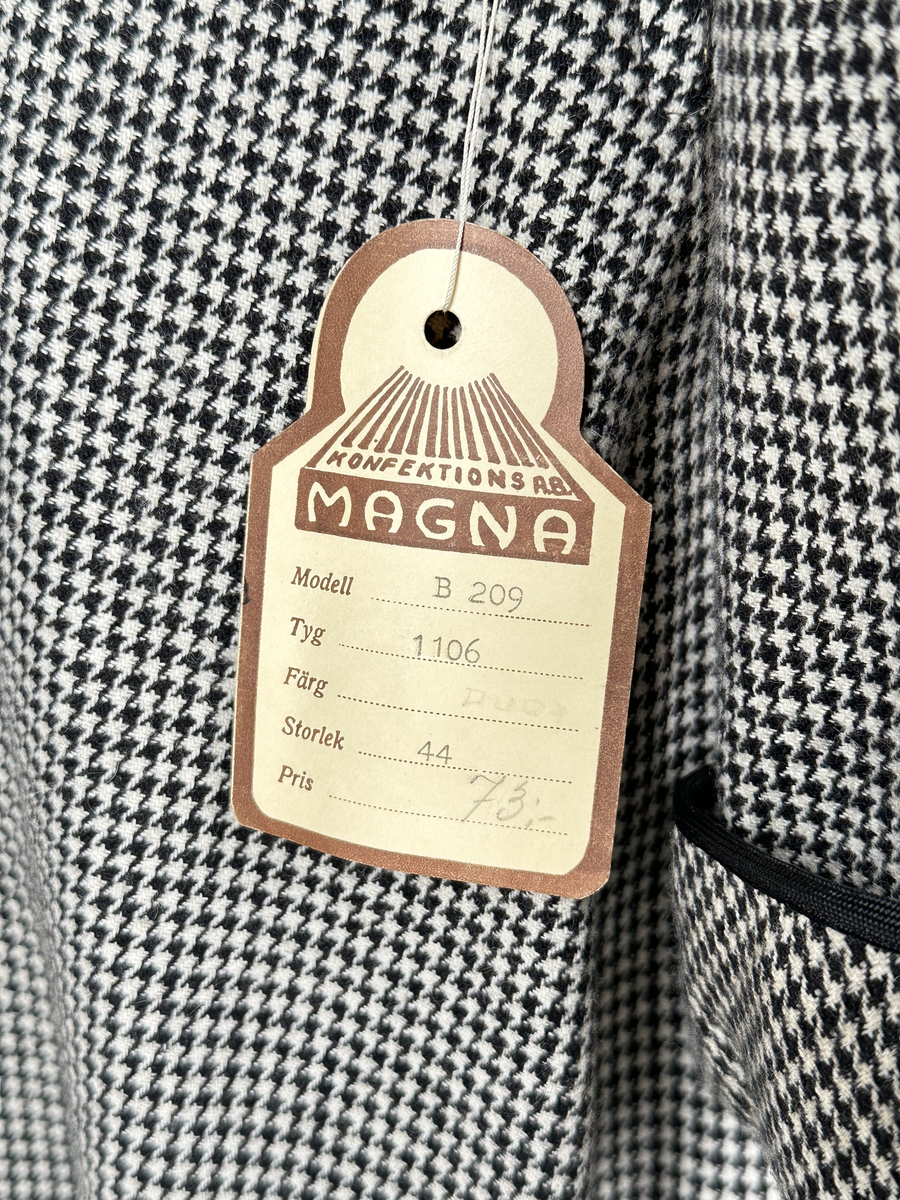 Klänning av hundtandsmönstrat ylletyg i svart och vitt. Krage och uppvikta manschetter kantade med svart band. Axelvaddar. Midjeskuren, med överdel som är öppen ner till midjan och försedd med isättning som är fäst i midjesömmen. Isättningen med dubbla knapprader och en tryckknapp överst, som knäppes till överdelen för att sluta öppningen. Tunna hällor för skärp på var sida om midjan. Dragkedja i sidan, under armhålan.