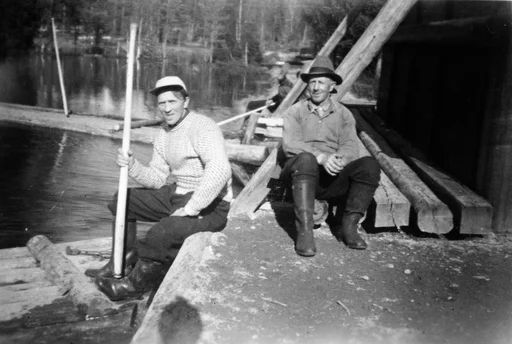 Fløting. To menn sitter på fløteinnretning, den ene med fløterhake, og beina på en flåte.