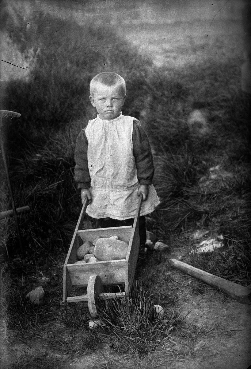 Barn med leketrillebår, triller stein. Gutt i kjole.