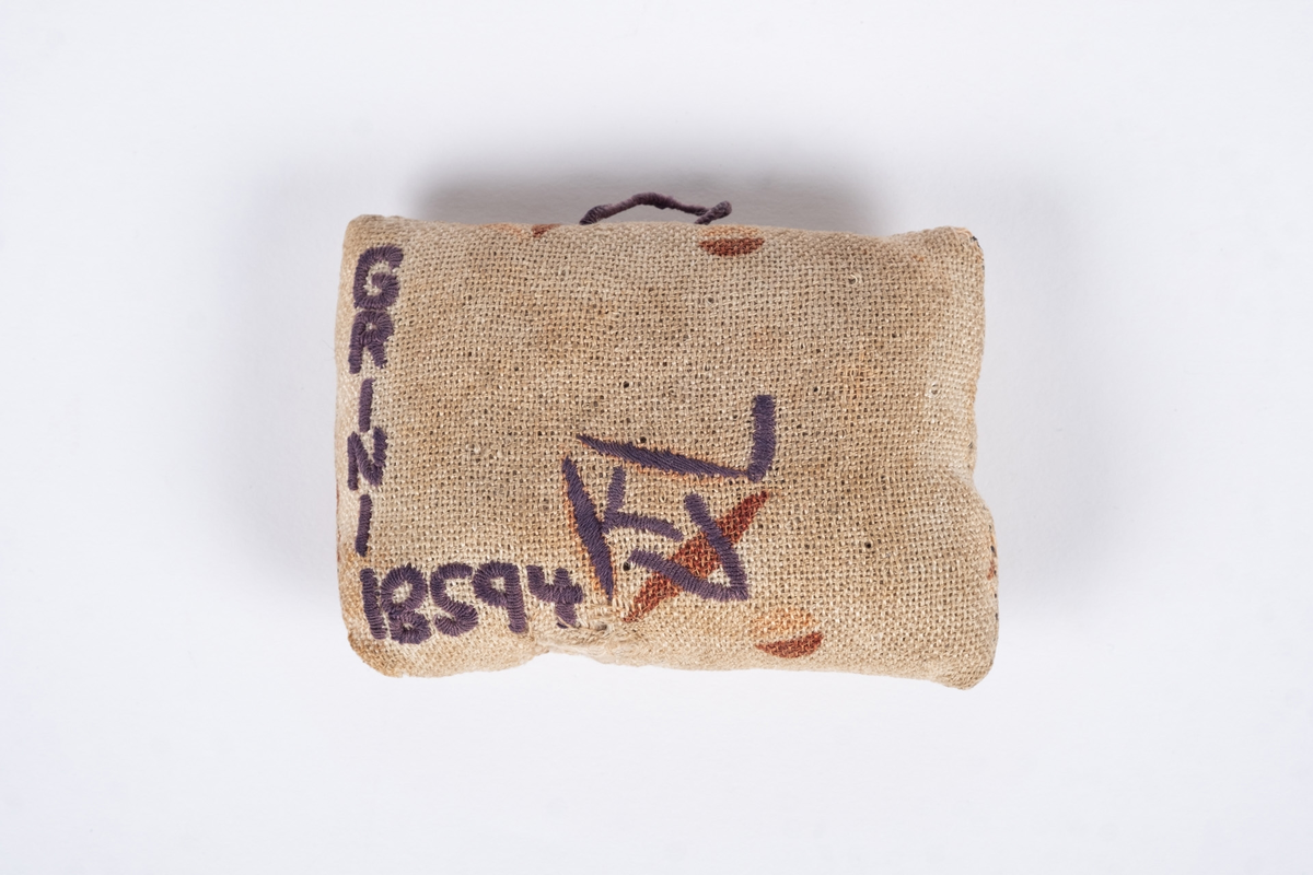 Liten nålepute sydd i grovt naturfarget lerretstoff, med geometrisk mønster i brunt og lyserødt.. Brodert tekst, fangenummer og datoer på for- og bakside. 