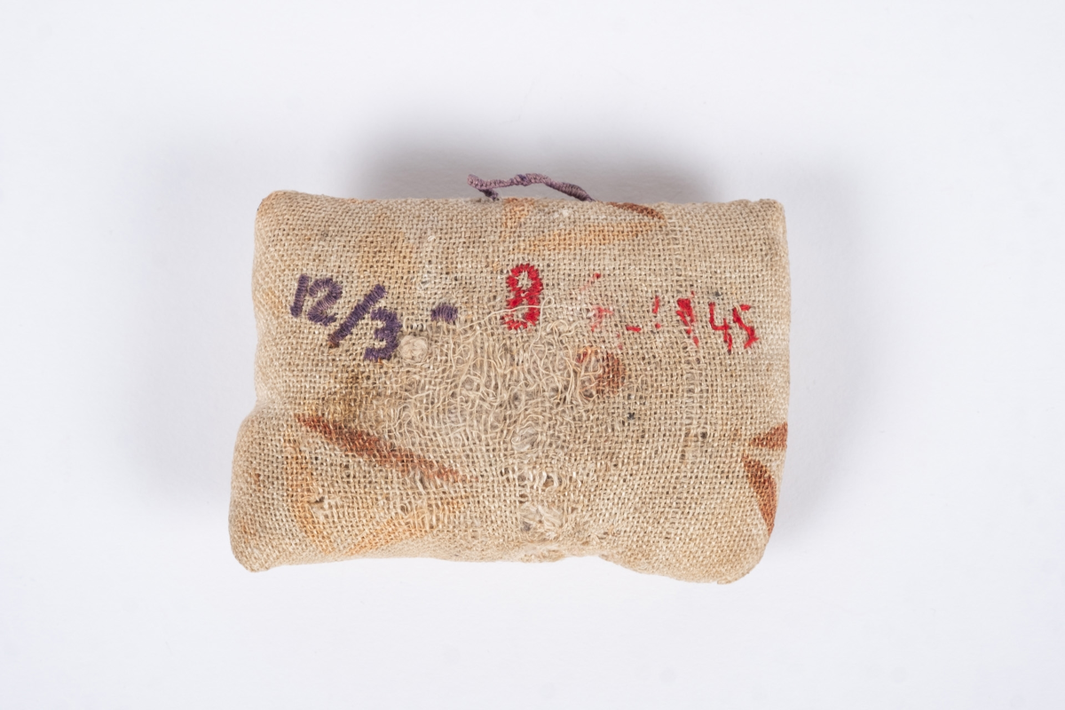 Liten nålepute sydd i grovt naturfarget lerretstoff, med geometrisk mønster i brunt og lyserødt.. Brodert tekst, fangenummer og datoer på for- og bakside. 