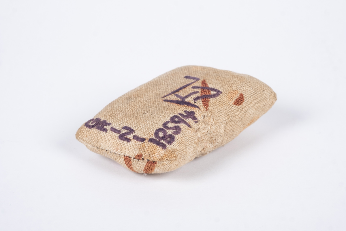 Liten nålepute sydd i grovt naturfarget lerretstoff, med geometrisk mønster i brunt og lyserødt.. Brodert tekst, fangenummer og datoer på for- og bakside. 