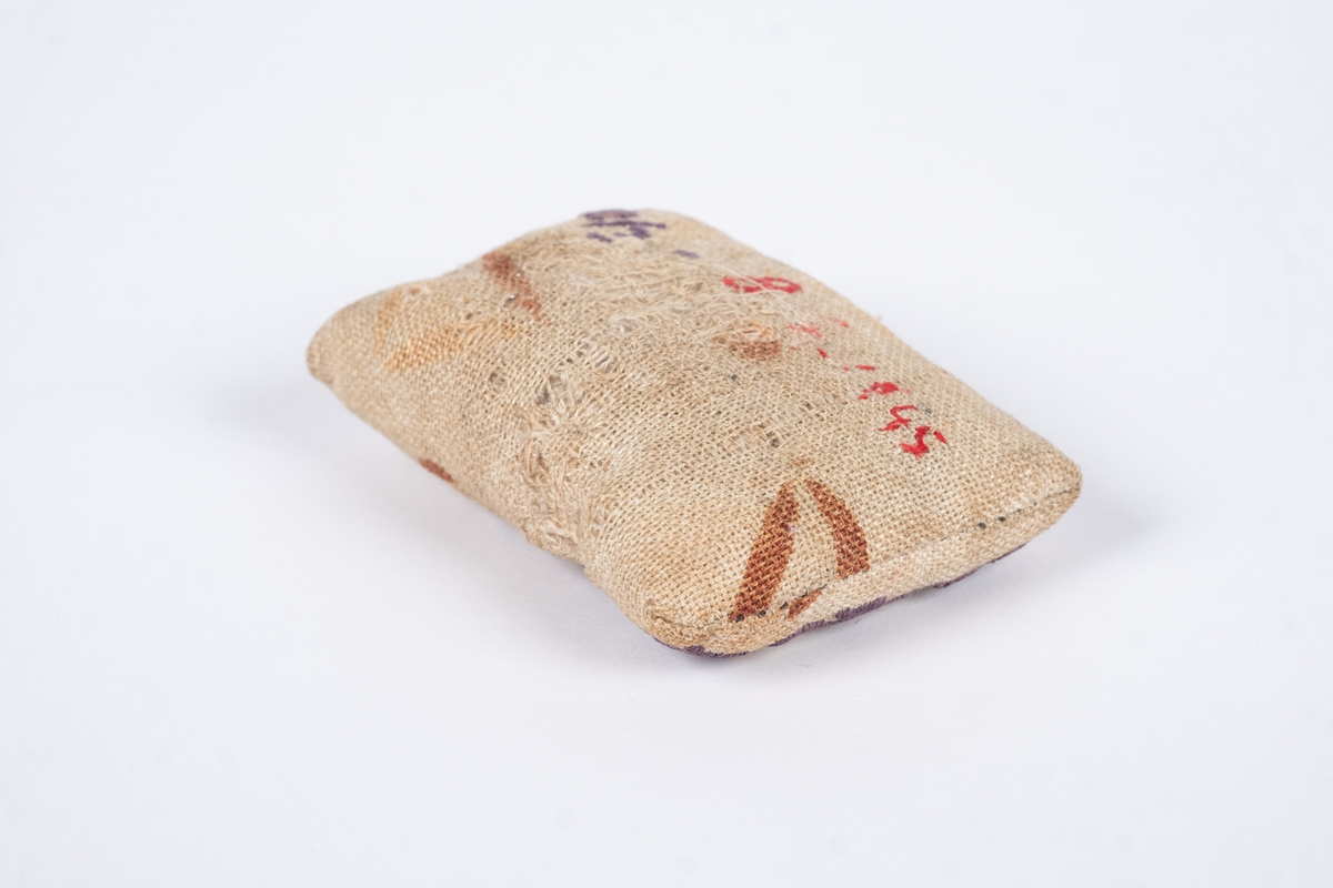 Liten nålepute sydd i grovt naturfarget lerretstoff, med geometrisk mønster i brunt og lyserødt.. Brodert tekst, fangenummer og datoer på for- og bakside. 