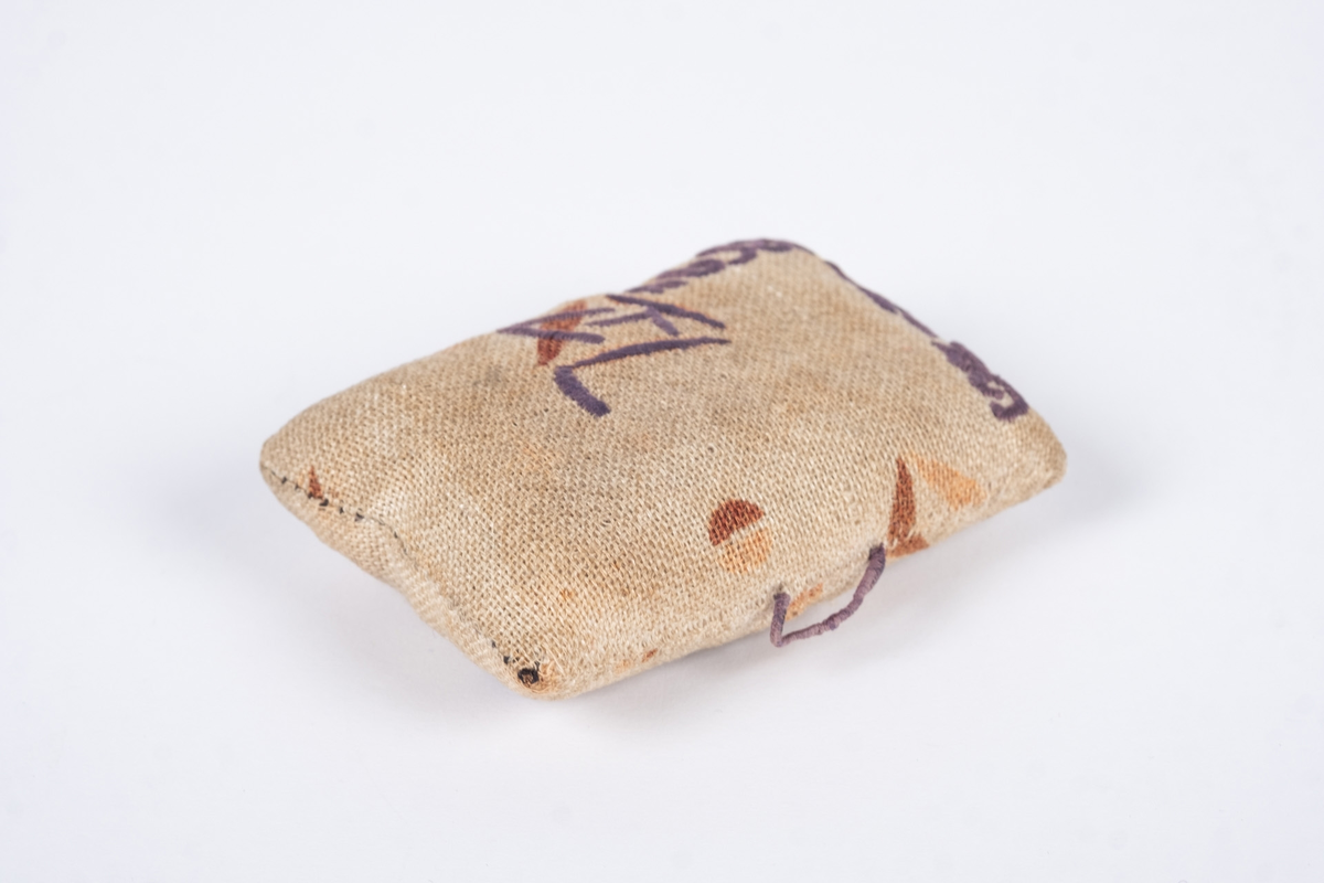 Liten nålepute sydd i grovt naturfarget lerretstoff, med geometrisk mønster i brunt og lyserødt.. Brodert tekst, fangenummer og datoer på for- og bakside. 