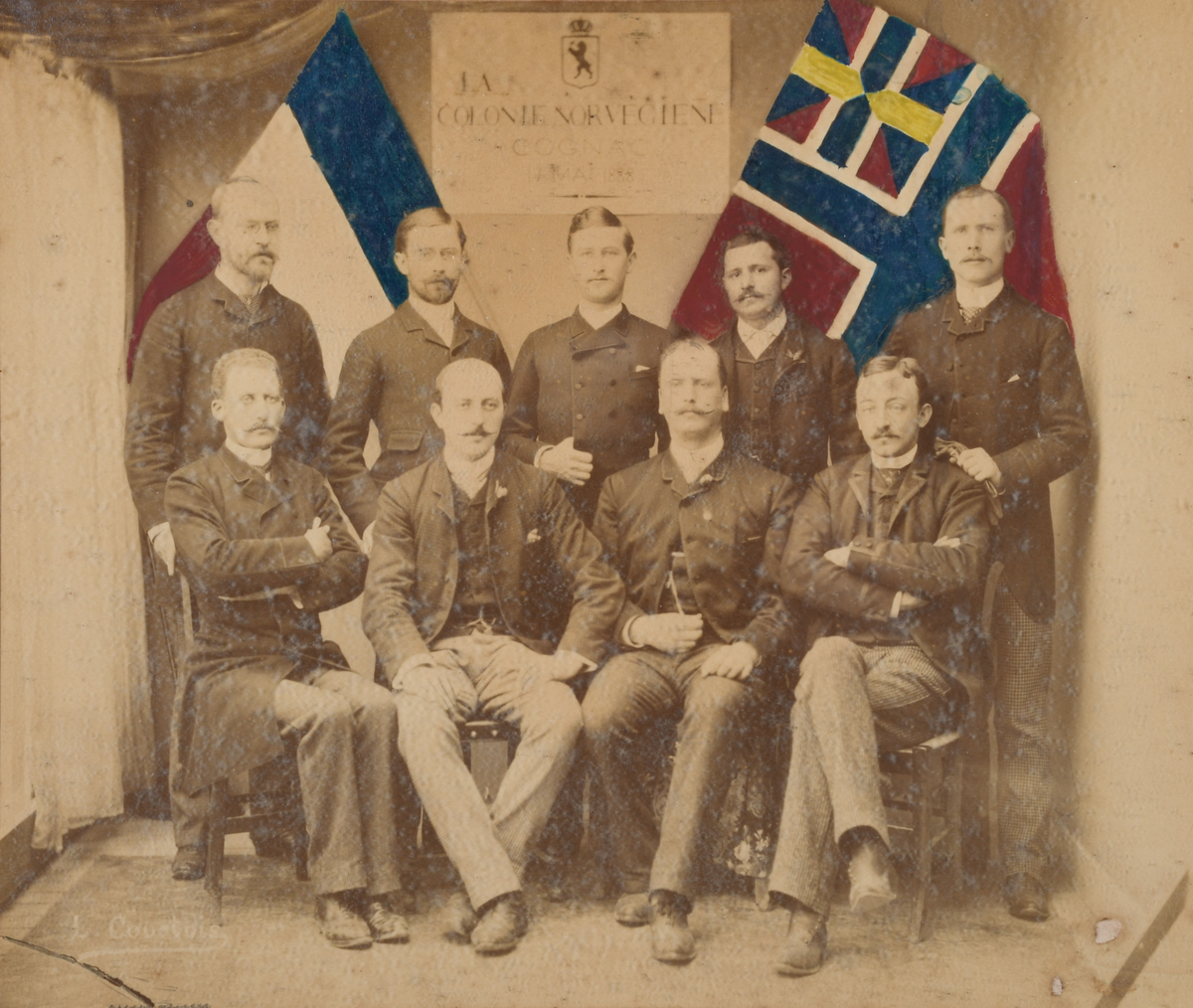 Gruppefotografi av ni menn, norskt handelsflagg og fransk nasjonalflagg i bakgrunnen. Ni signaturer under fotografiet.