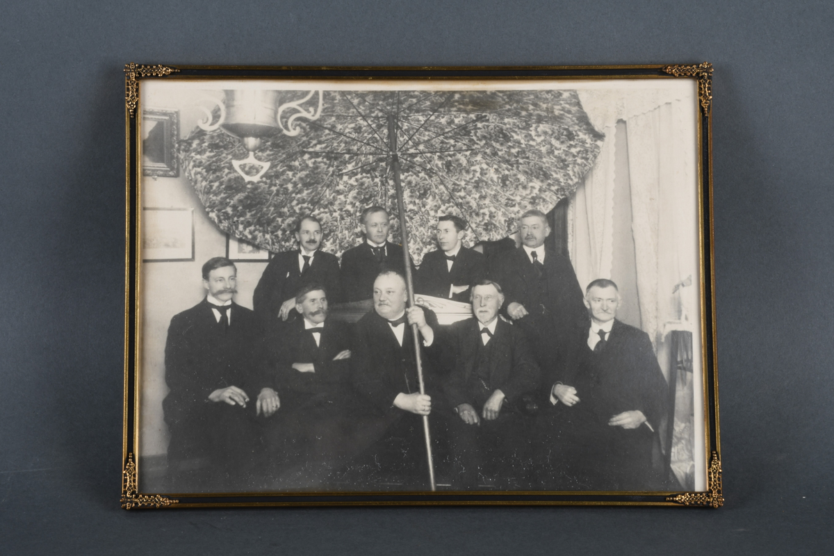 Innrammet svart-hvitt foto med rektangulær form. Motivet viser ni menn kledd i dress, stående og sittende i en stue. Mannen foran i midten holder en stor parasol. Rammen er av forgylt og svart metall med dekor i hjørnene, samt beskyttende, konvekst glass.