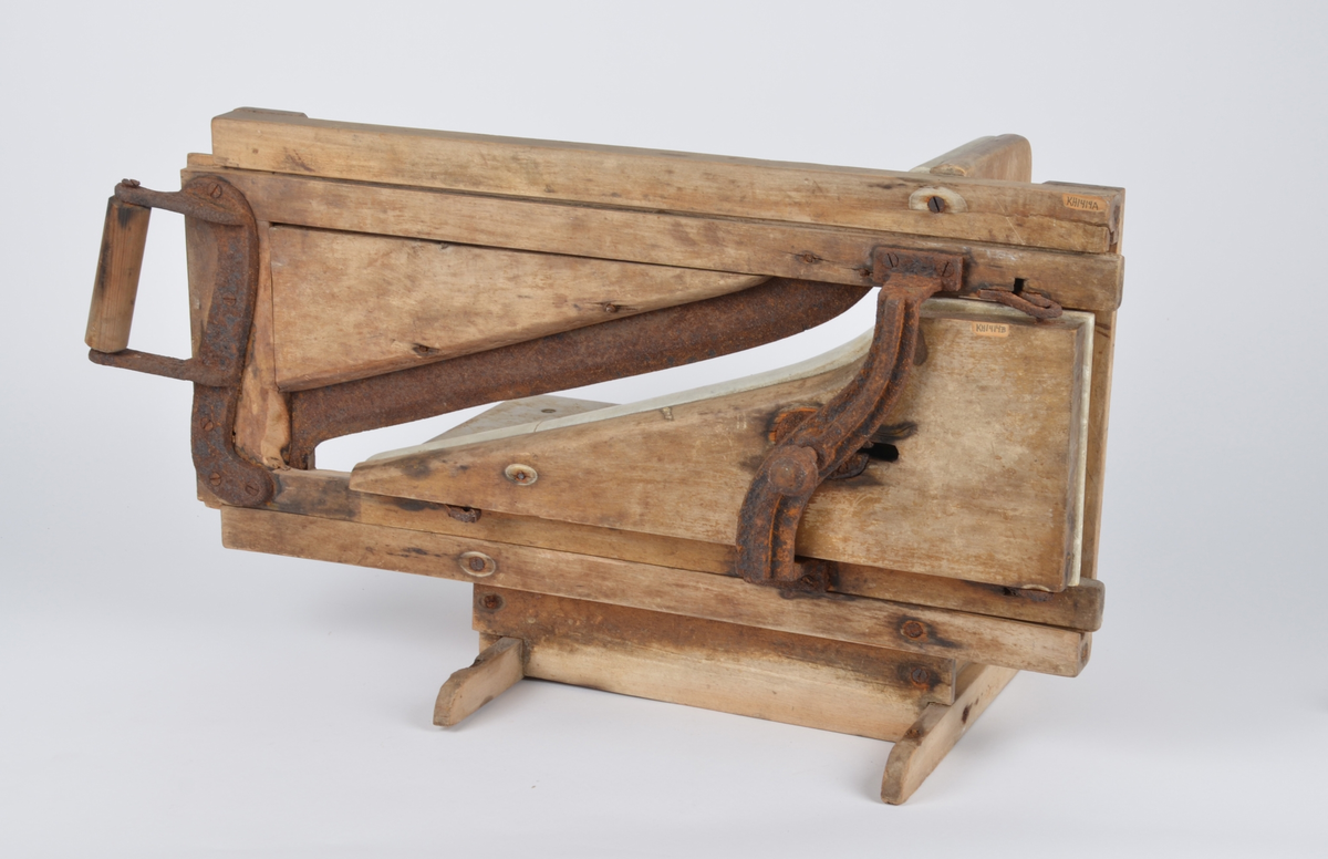 Påleggskjæremaskin av tre, metall og marmor. Base av tre med to føtter. Skjærefjøl av marmor med bakplate. På ene siden en ramme til knivslede. Løs knivslede med sideplate av marmor og håndtak.
