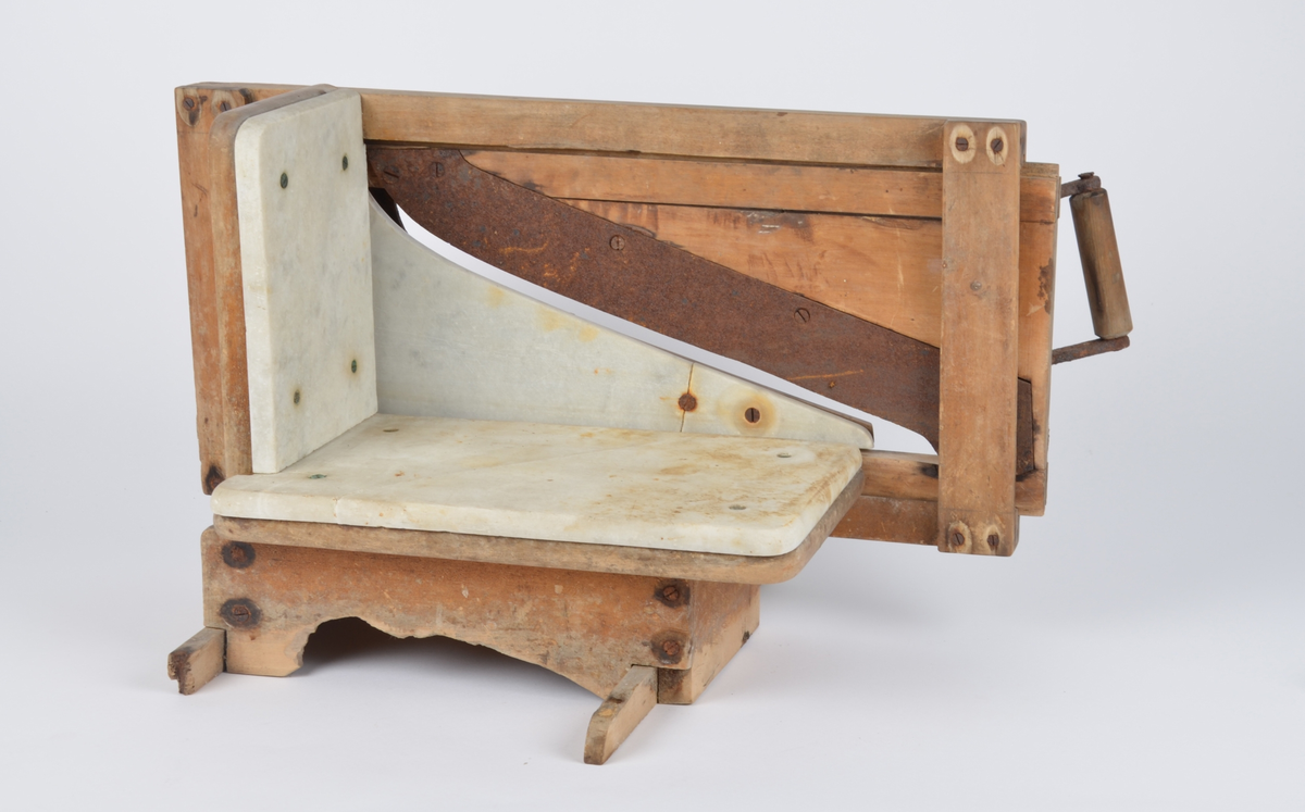 Påleggskjæremaskin av tre, metall og marmor. Base av tre med to føtter. Skjærefjøl av marmor med bakplate. På ene siden en ramme til knivslede. Løs knivslede med sideplate av marmor og håndtak.