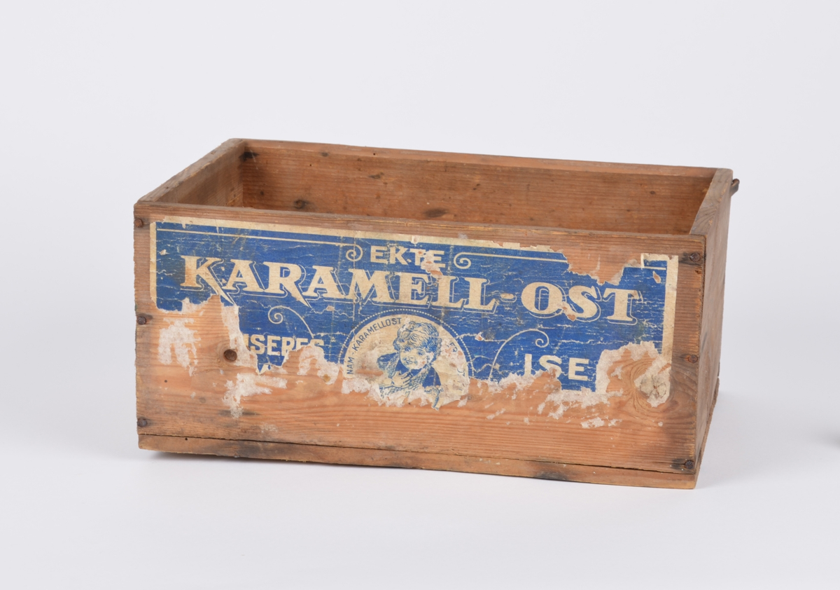 Rektangulær trekasse. På ene langsiden pålimt papiretikett i hvitt og blått med reklame for karamellost.