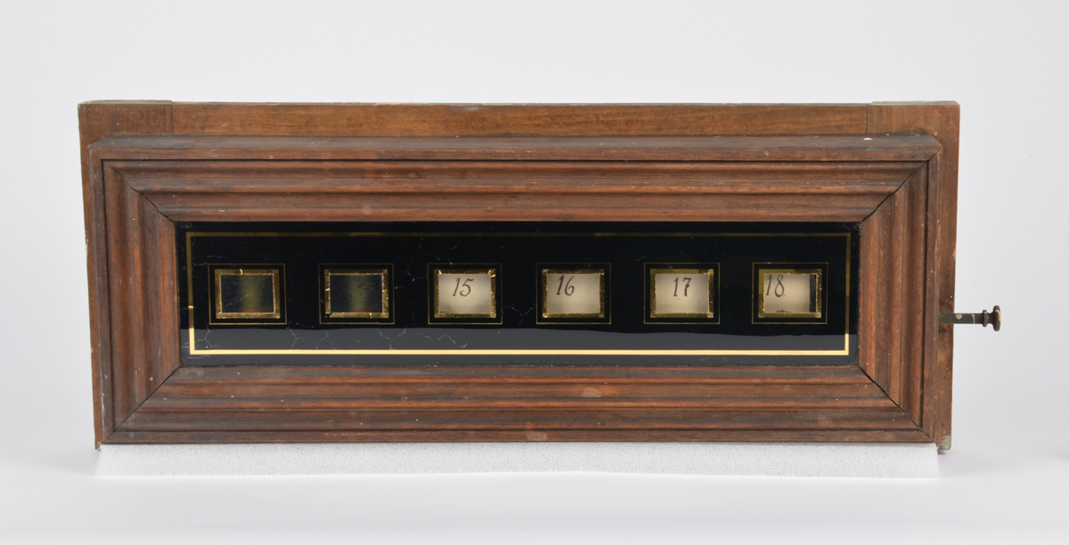 Rektangulær trekasse. I midten et svartmalt glasspanel med gjennomsiktige felter hvor brikker med nummer vises. På ene kortsiden av kassen en knapp som kan trykkes inn for å nullstille nummerbrikkene