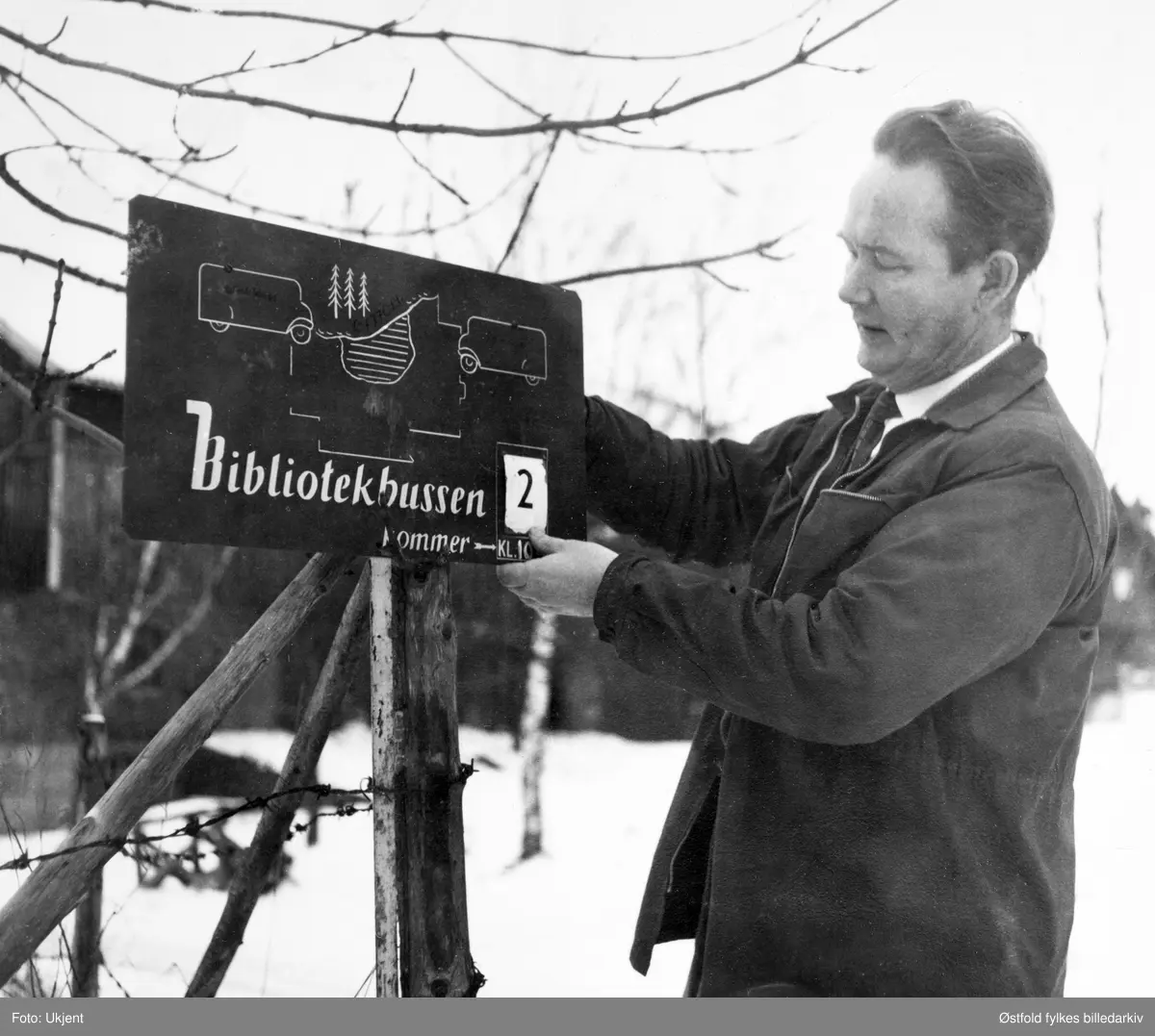 Bokbussen fra Østfold sentralbibliotek i Fredrikstad besøker Varteig i desember 1960. Sjåfør Viggo Nilsen fra Østfold sentralbibliotek i Fredrikstad henger opp nytt datoskilt på en av de faste oppslagsplakatene for bokbussens ankomsttid  (som var laget etter amerikansk system).  
I 1960 hadde bokbussen 7000 låntakere og besøkte 122 steder i Østfold.  10.-15.000 bøker sirkulerte i bygdene der bokbussen kjørte, og bussen hadde med seg ca.  2000 bøker i  bussen,  Folk møtte opp til faste holdeplasser på faste dager, hver fjerde uke. Det var også mulig å bestille bøker. Om sommeren var bokbussens ruter innstilt.
Viggo Nilsen startet som sjåfør for bokbussen ca. 1951-52., og kjørte hver eneste rute, mens bibliotekarene i Fredrikstad skiftet på å være med. Nilsen arbeidet som bokbussjåfør til han ble pensjonist i 1984.
Skilt: "Bokbussen kommer", se Avbildet obkekt.
