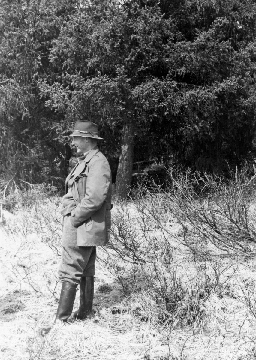 Nils Sand (1908-1988), som var fløtersjef i elva Vidmunda, fotografert under ei befaring langs dette vassdraget i 1958. Fløtersjefen var kledd i vadmelsbuker og ei dobbeltspent vindjakke, som da bildet ble tatt var ukneppet. På hodet hadde han en litt slitt filthatt, og på beina langskaftete gummistøvler. Da bildet ble tatt smilte han. Bildet er antakelig tatt like ved vassdraget, der det vokste vierkratt med granskog i bakgrunnen.