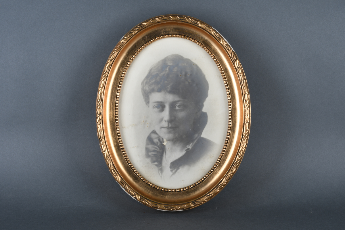 Innrammet svart-hvitt foto med oval form. Motivet viser portrett av en kvinne som ser forbi kamera. Hun har høy krage og oppsatt hår. Rammen er gullforgylt med dekorativ bord ytterst og perlebord innerst, samt beskyttende glass.