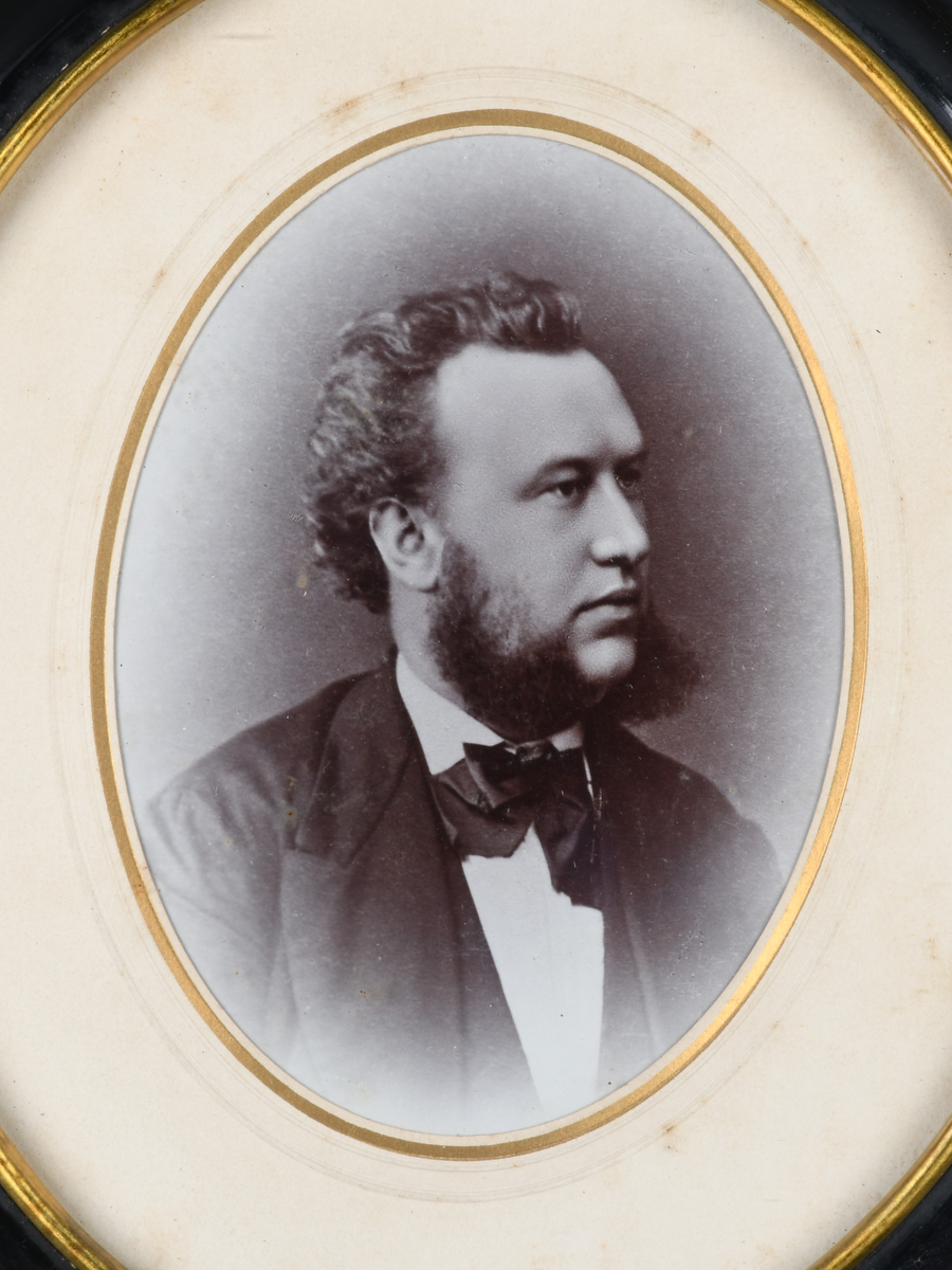 Innrammet svart-hvitt foto med oval form. Motivet viser portrett av en mann som ser til siden. Han har skjegg, hvit skjorte og svart jakke, vest og sløyfe. Foto har passepartout av kartong med riller og forgylt kant innerst. Rammen er svartmalt og profilert med metallkant innerst, samt beskyttende glass.