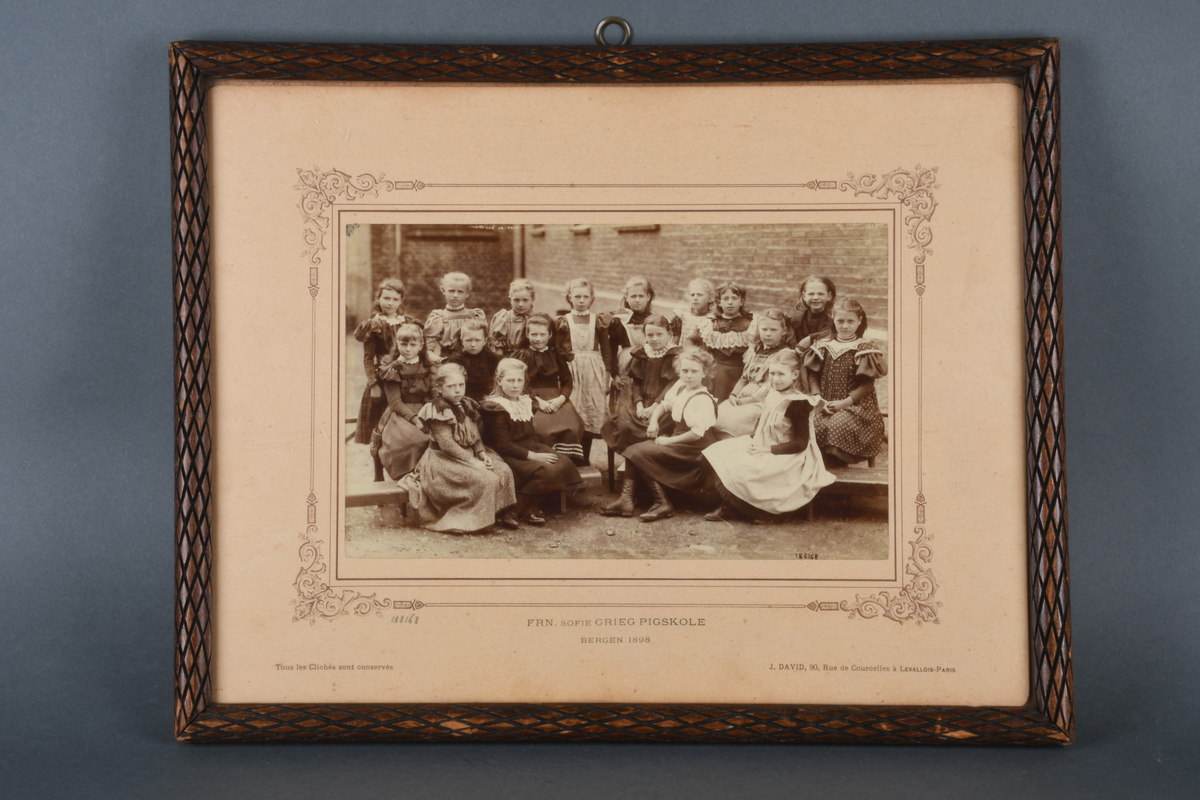 Innrammet foto med rektangulær form. Motivet viser 18 pikebarn stående og sittende utenfor en murbygning. Foto er limt på en beige kartong med bord med dekorative hjørner rundt foto. Det er rammet inn i en brun treramme med utskjært rutemønster, samt beskyttende glass.