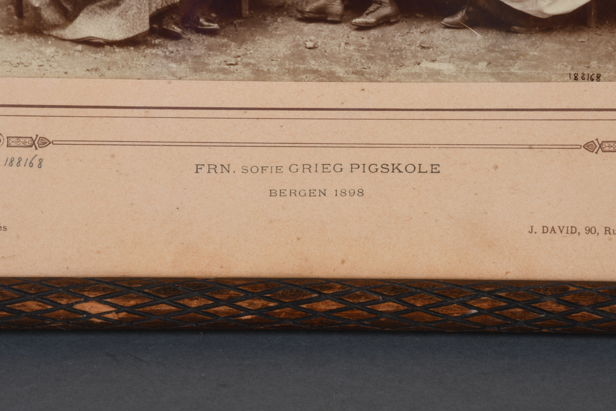 Innrammet foto med rektangulær form. Motivet viser 18 pikebarn stående og sittende utenfor en murbygning. Foto er limt på en beige kartong med bord med dekorative hjørner rundt foto. Det er rammet inn i en brun treramme med utskjært rutemønster, samt beskyttende glass.