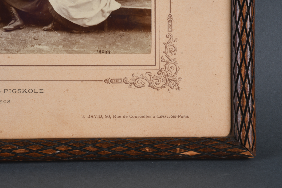 Innrammet foto med rektangulær form. Motivet viser 18 pikebarn stående og sittende utenfor en murbygning. Foto er limt på en beige kartong med bord med dekorative hjørner rundt foto. Det er rammet inn i en brun treramme med utskjært rutemønster, samt beskyttende glass.