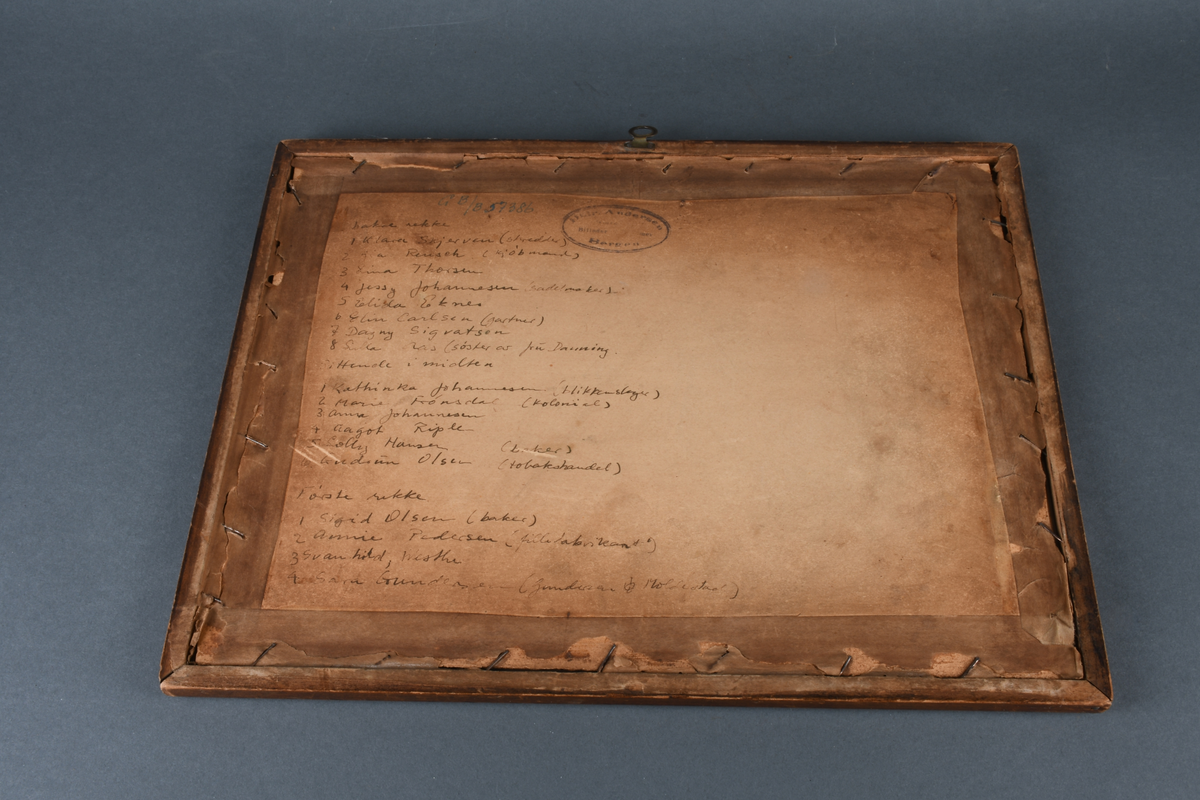 Innrammet foto med rektangulær form. Motivet viser 18 pikebarn stående og sittende utenfor en murbygning. Foto er limt på en beige kartong med bord med dekorative hjørner rundt foto. Det er rammet inn i en brun treramme med utskjært rutemønster, samt beskyttende glass.