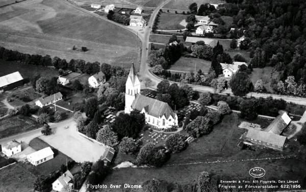 Vykort, "Flygfoto över Okome." Okome kyrka och kyrkogård med omgivade bebyggelse vid trevägskorsningen mellan Ätrafors, Ullared och Gällared. Prästgården ligger närmast till höger om kyrkan. Villabebyggelse. Vid den bortre vägskorsningen ligger affären och mitt för den Okome skola.