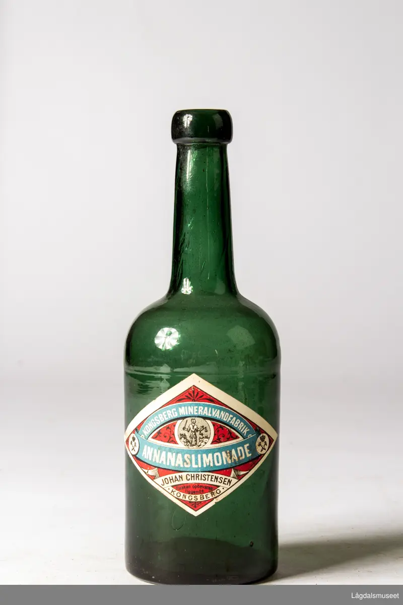 Grønn glassflaske med etikett. Etiketten har Kongsbergs gamle kommunevåpen og bergmannssymbolet korslagt hammer og bergsjern. Det siste med krone over. 
