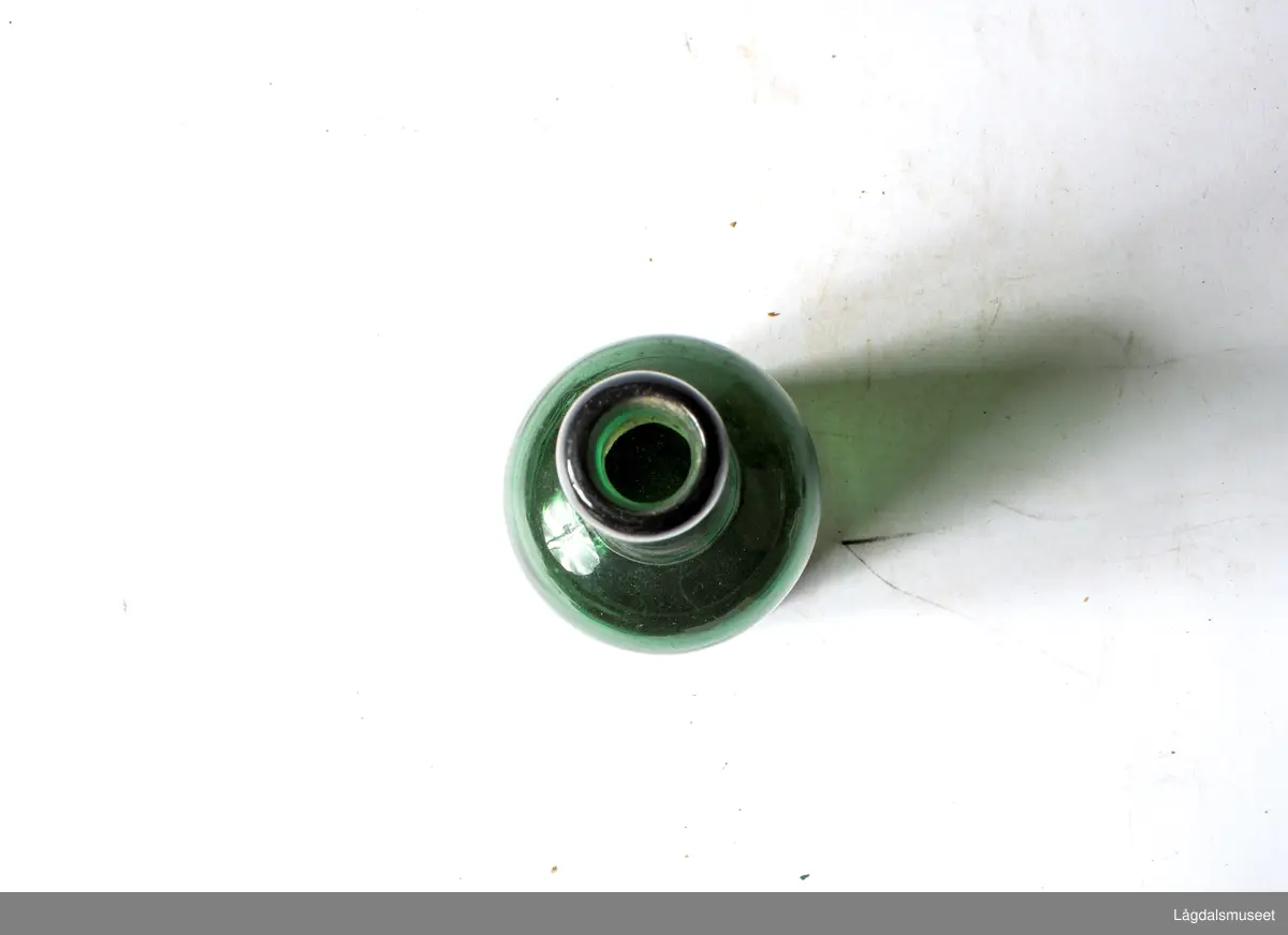 Grønn glassflaske med etikett. Etiketten har Kongsbergs gamle kommunevåpen og bergmannssymbolet korslagt hammer og bergsjern. Det siste med krone over. 