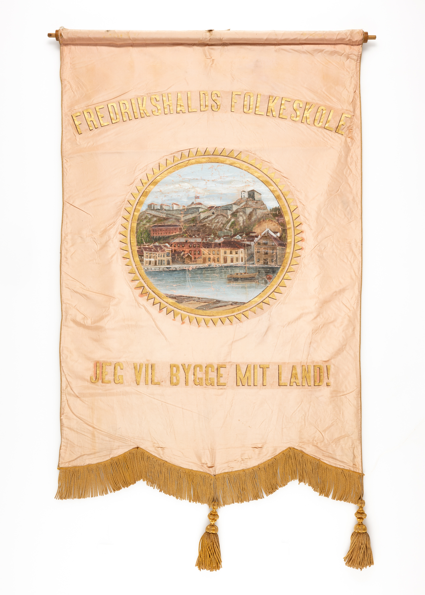 Ensidig i fane rosa og kremgul silke med malt motiv. Løpegang i øvre del. Råstang av tre, mangler sidespyd, snorer etc. Frynser i trefliket nederkant. FORSIDE: Midtmotiv i tindet sirkel, flerfarget: Fredriksten festning med bebyggelse på Kongens Brygge i forgrunnen. Båt på sjøen. Tekst over motiv i gullskrift og i svak bue: FREDRIKSHALDS FOLKESKOLE. Tekst under motiv: JEG VIL BYGGE MIT LAND! BAKSIDE: Kremgul uten tekst, dekor eller motiv.