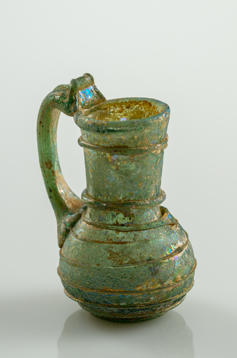 Liten kanna, med grep, i gröntonat glas. Pålagd glastråd som ringlar sig från botten till mynningen.