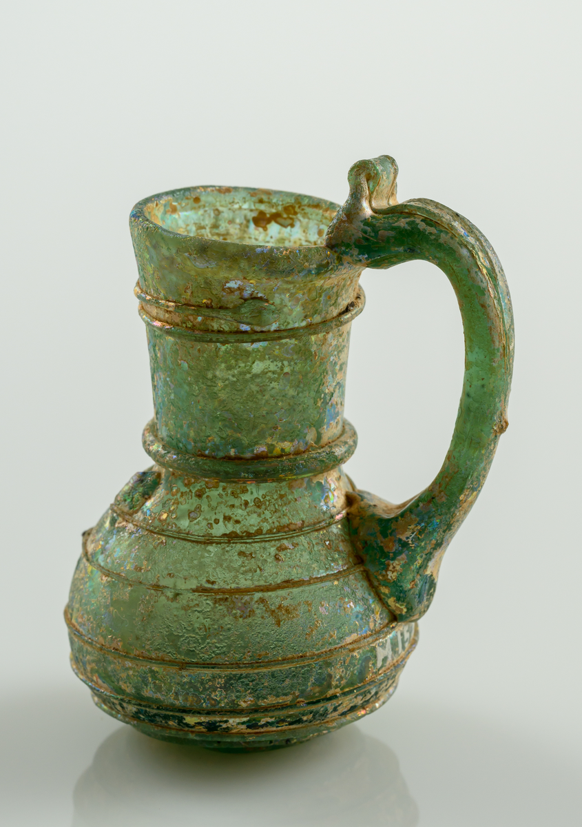 Liten kanna, med grep, i gröntonat glas. Pålagd glastråd som ringlar sig från botten till mynningen.