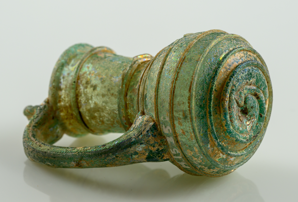 Liten kanna, med grep, i gröntonat glas. Pålagd glastråd som ringlar sig från botten till mynningen.