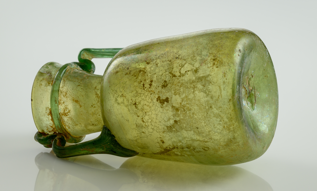 Urna i gröntonat glas, med dubbla grepar. Svagt konisk nedre del som övergår i en bred hals som vidgar sig uppåt.