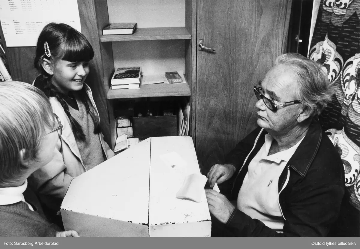 Bokbussen fra Østfold fylkesbibliotek i Rakkestad  26.  august 1982. Bokbussjåfør og bibliotekfullmektig Viggo Nilsen skriver ut lånekort til nye lesehester (ukjente) som besøker bokbussen. Barna er fra Ytterskogen skole i Rakkestad.
