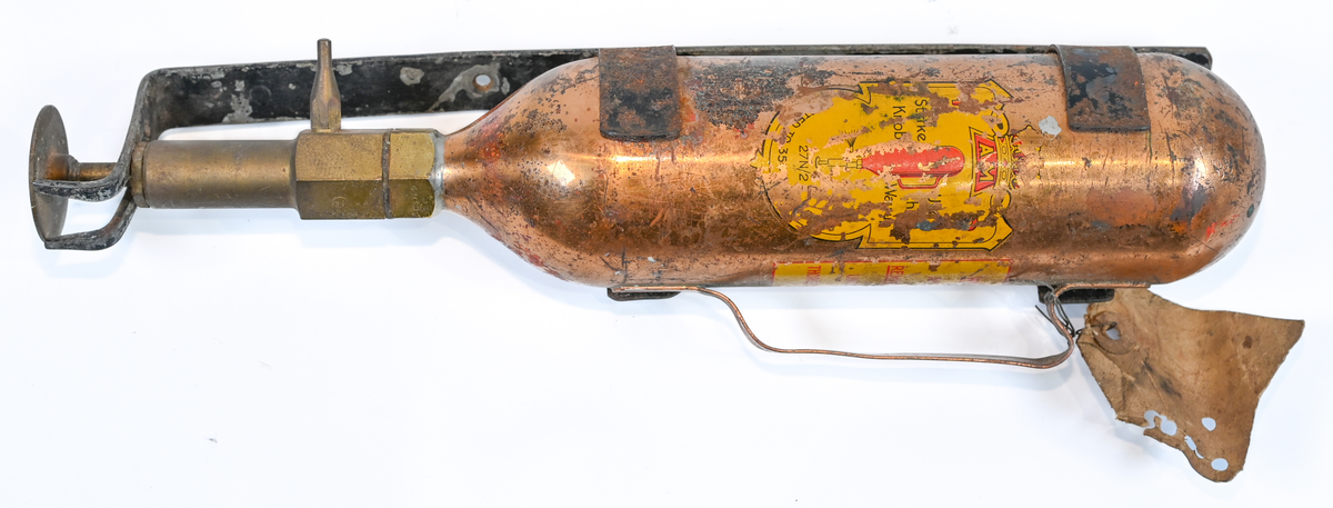 Brandsläckare i röd metall med väggstativ i järn. Brandsläckaren utlöstes genom att slå den nedre ventilen i marken. Väggstativet utgör en säkerhet för att undvika oönskade aktivering av brandsläckaren. På sidan av och runt om föremålet ser man rester av en etikett med engelsk text som agerar som instruktioner för användning. Texten är svår att läsa. På föremålet hittades en etikett med svensk text, okänt ursprung.