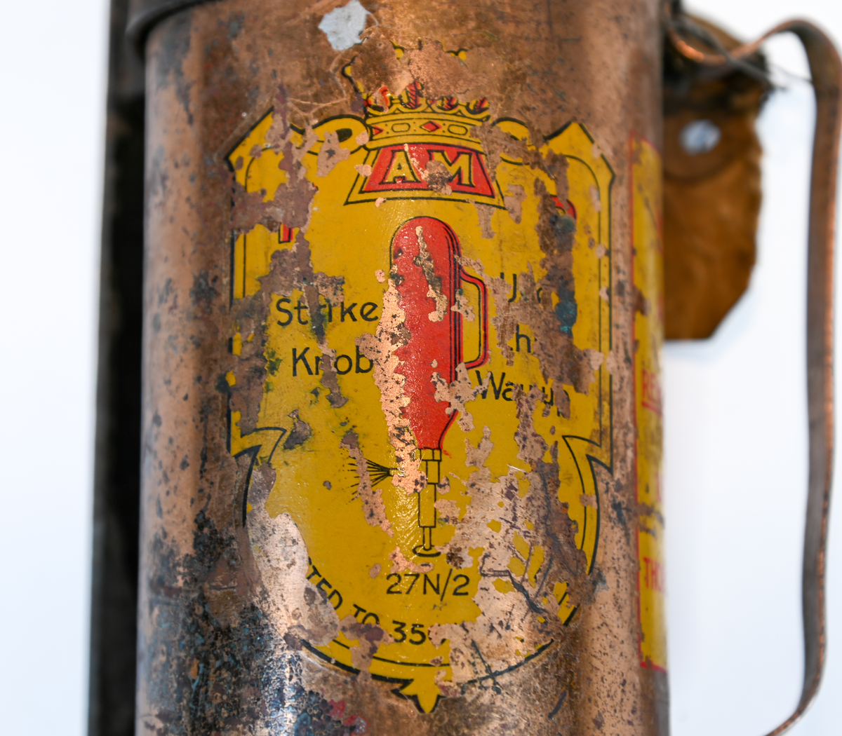 Brandsläckare i röd metall med väggstativ i järn. Brandsläckaren utlöstes genom att slå den nedre ventilen i marken. Väggstativet utgör en säkerhet för att undvika oönskade aktivering av brandsläckaren. På sidan av och runt om föremålet ser man rester av en etikett med engelsk text som agerar som instruktioner för användning. Texten är svår att läsa. På föremålet hittades en etikett med svensk text, okänt ursprung.