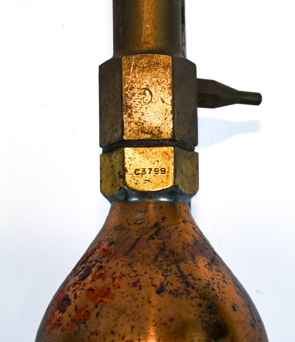 Brandsläckare i röd metall med väggstativ i järn. Brandsläckaren utlöstes genom att slå den nedre ventilen i marken. Väggstativet utgör en säkerhet för att undvika oönskade aktivering av brandsläckaren. På sidan av och runt om föremålet ser man rester av en etikett med engelsk text som agerar som instruktioner för användning. Texten är svår att läsa. På föremålet hittades en etikett med svensk text, okänt ursprung.