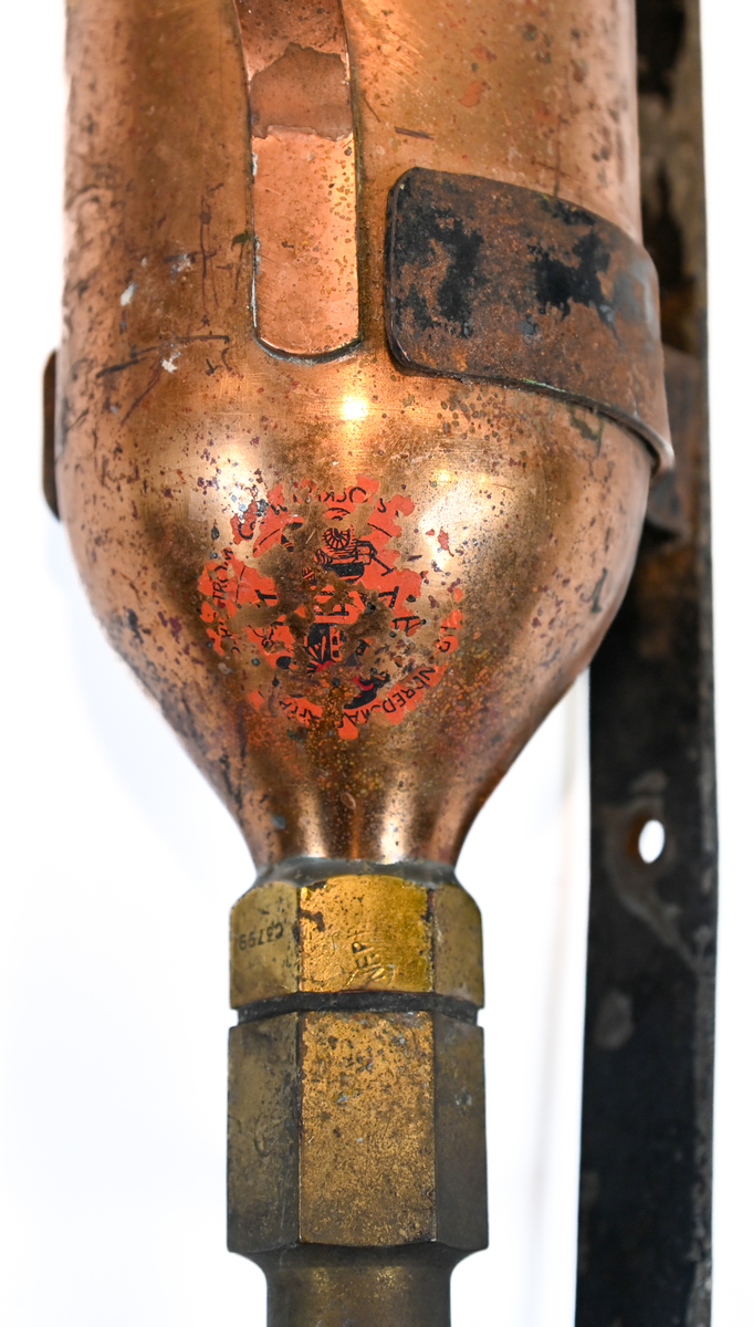 Brandsläckare i röd metall med väggstativ i järn. Brandsläckaren utlöstes genom att slå den nedre ventilen i marken. Väggstativet utgör en säkerhet för att undvika oönskade aktivering av brandsläckaren. På sidan av och runt om föremålet ser man rester av en etikett med engelsk text som agerar som instruktioner för användning. Texten är svår att läsa. På föremålet hittades en etikett med svensk text, okänt ursprung.