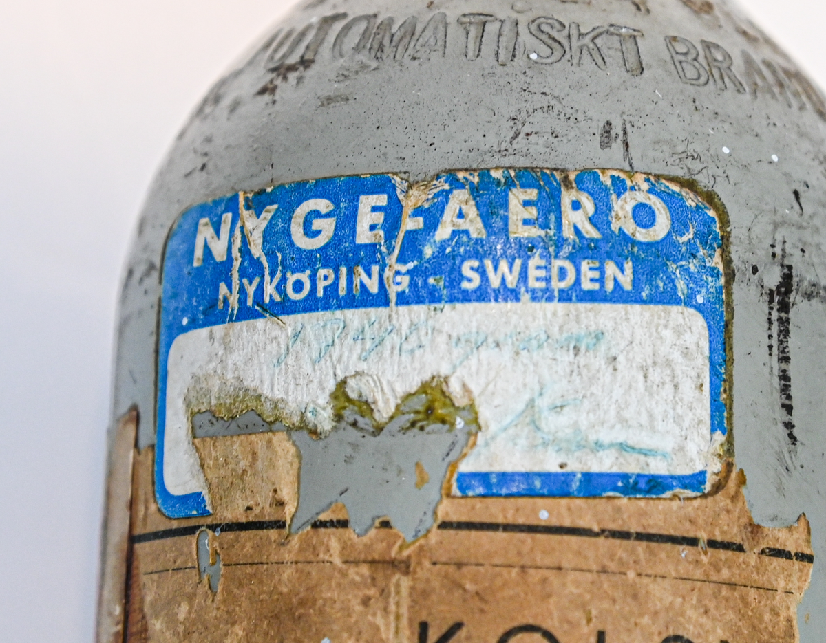 Kolsyreflaska i silverfärgad metall, förmodligen ansett som en del av en brandsläckare system. Flaskan är märkt som tillverkad av De Förenade Kolsyrefabrikernas AB 1/3/1952 och med sista inspektion av Swedish Airworks LTD 27/02/1974. På sidan finns det instruktion för hantering av föremålet som gasflaska på ena sidan och som brandsläckare på andra sidan. Fylld vikt: 1.31 kg. Prägling som attesterat att flaskan fick inspektion 1958, 1965 och 1972.