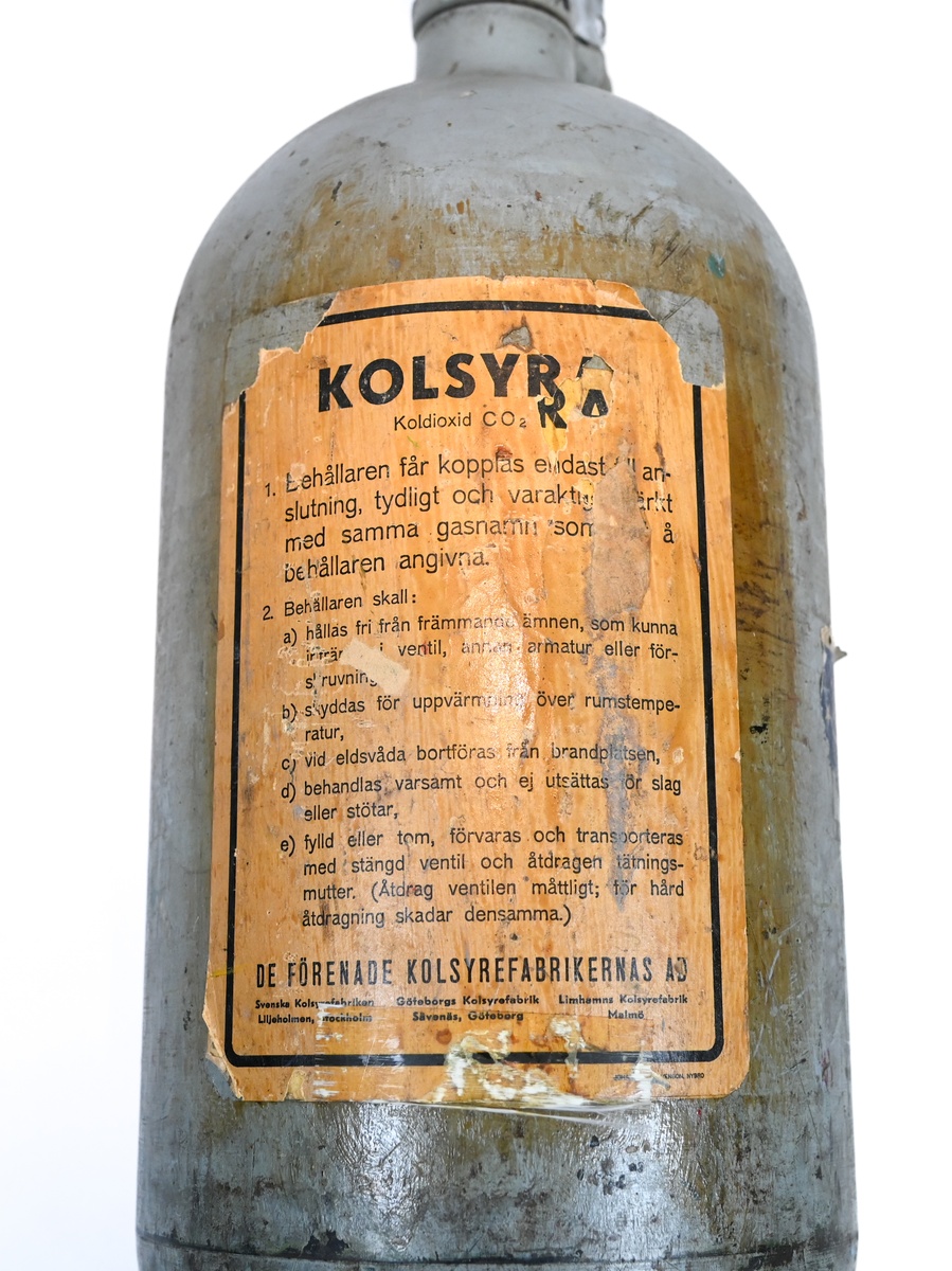 Kolsyresläckare i gråfärgad avrundad metall flaska med ventil, lösningshandtag och slang i fiber och läder, tillverkad av De Förenade Kolsyrefabrikernas AB. På föremålet står det ABA Typ-501-32-02-1. Tillsynsdatum präglat på flaskan 10/40 och 07/46.