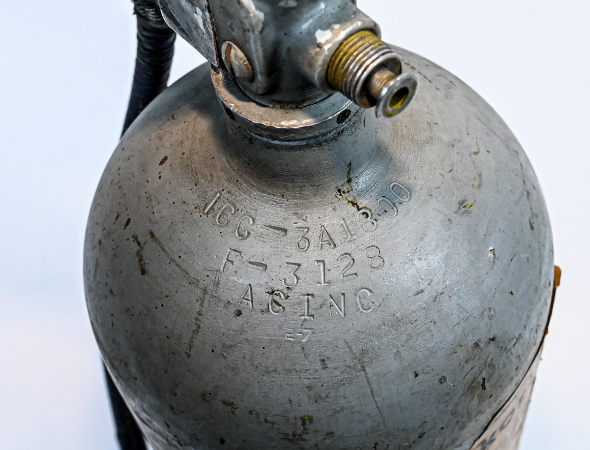 Kolsyresläckare i gråfärgad avrundad metall flaska med ventil, lösningshandtag och slang i fiber och läder, tillverkad av De Förenade Kolsyrefabrikernas AB. På föremålet står det ABA Typ-501-32-02-1. Tillsynsdatum präglat på flaskan 10/40 och 07/46.