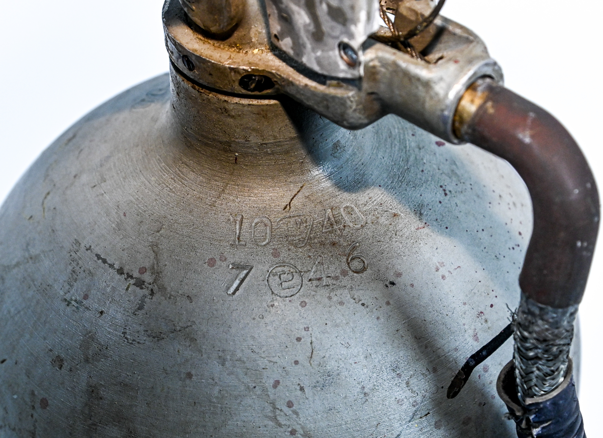 Kolsyresläckare i gråfärgad avrundad metall flaska med ventil, lösningshandtag och slang i fiber och läder, tillverkad av De Förenade Kolsyrefabrikernas AB. På föremålet står det ABA Typ-501-32-02-1. Tillsynsdatum präglat på flaskan 10/40 och 07/46.