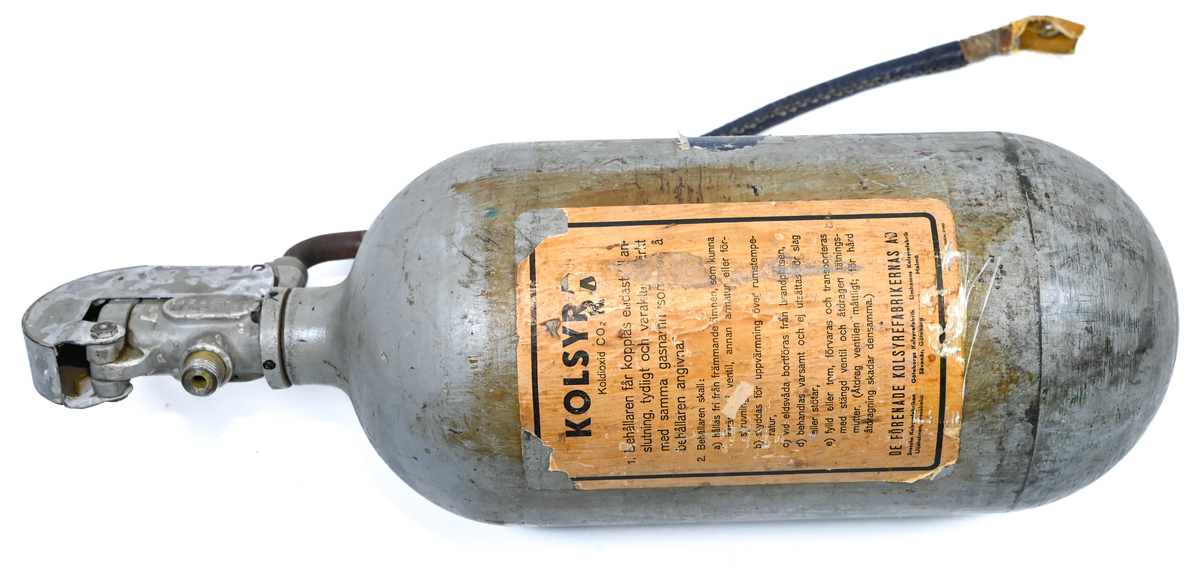 Kolsyresläckare i gråfärgad avrundad metall flaska med ventil, lösningshandtag och slang i fiber och läder, tillverkad av De Förenade Kolsyrefabrikernas AB. På föremålet står det ABA Typ-501-32-02-1. Tillsynsdatum präglat på flaskan 10/40 och 07/46.
