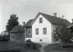 Här ser vi bostadshuset från trädgården. Huset har ett bakbygge i trä innehållande kök och ett bakbygge på bakbygget innehållande brygghus (?). Man har klätt huset och köksbakbygget med papp, medan det yttersta bakbygget ännu har spåntak.