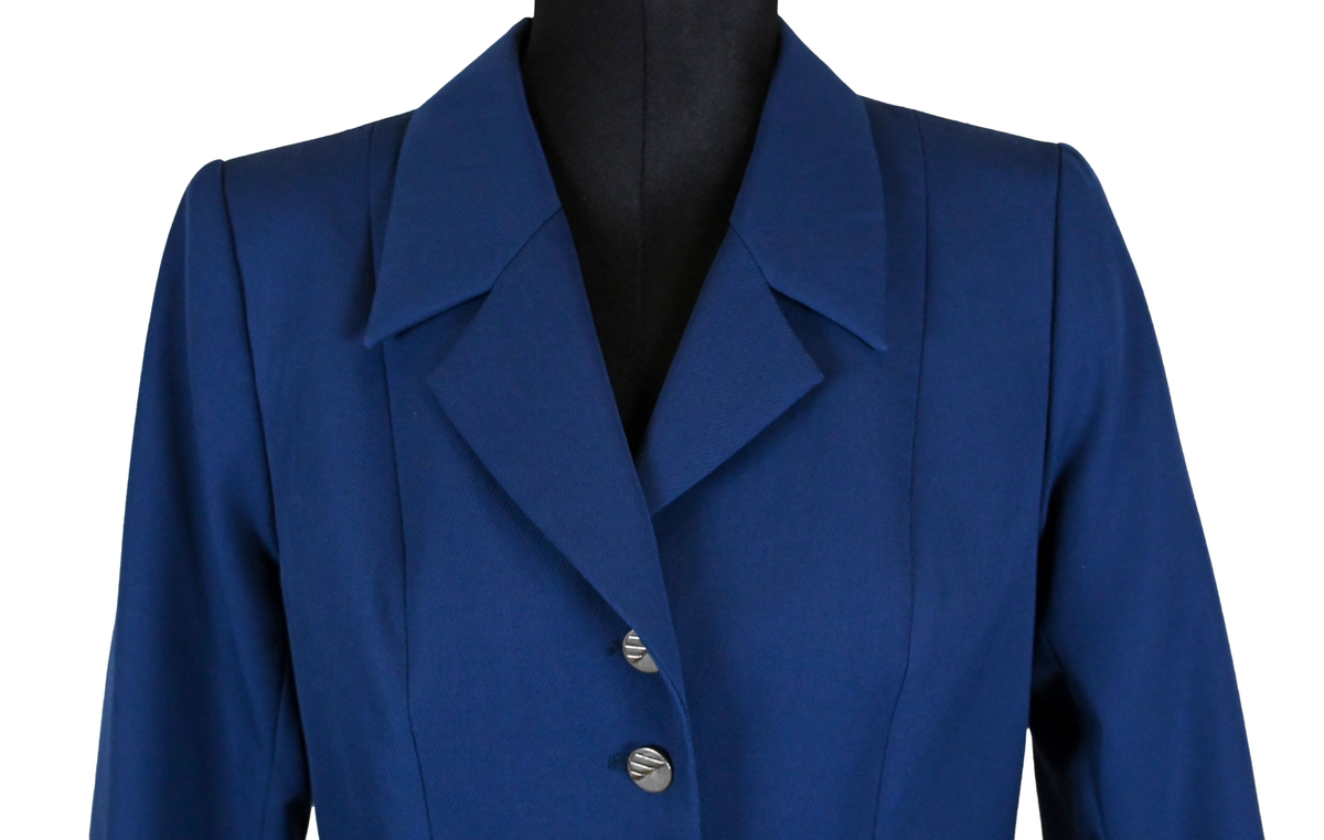 Kavaj i mörklbå ull- och polysterblandning till ämbetsdräkt för kvinnlig personal vid Luftfartsverket. Fodret är gjort i mörkblå polyester. Kavajen har två framfickor med lock som fortfarande är igensydda samt en innerficka vänster sida som stängs med plastknapp. Kavajen stängs med metallknappar med Luftfartsverkets logga i relief. 

På insidan ovanför innerfickan sitter en lapp med Luftfartsverkets logga samt information om tillverkare och designer. I anslutnings till den sitter en mindre lapp med storleksangivelser. Kavajen producerad av Confetti och designad av Morgan i storlek C42. På insidan i ena sidosömmen sitter ytterligare en lapp med tvättanvisningar och information om material och tillverkningsort. Kavajan är tillverkad i Estland. I denna lapp sitter en extra metallknapp samt en fasthäftad papperslapp med numret "8632" i tryck och "3/10" påskrivet i blått bläck.