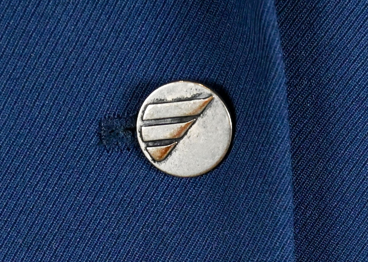 Kavaj i mörklbå ull- och polysterblandning till ämbetsdräkt för kvinnlig personal vid Luftfartsverket. Fodret är gjort i mörkblå polyester. Kavajen har två framfickor med lock som fortfarande är igensydda samt en innerficka vänster sida som stängs med plastknapp. Kavajen stängs med metallknappar med Luftfartsverkets logga i relief. 

På insidan ovanför innerfickan sitter en lapp med Luftfartsverkets logga samt information om tillverkare och designer. I anslutnings till den sitter en mindre lapp med storleksangivelser. Kavajen producerad av Confetti och designad av Morgan i storlek C42. På insidan i ena sidosömmen sitter ytterligare en lapp med tvättanvisningar och information om material och tillverkningsort. Kavajan är tillverkad i Estland. I denna lapp sitter en extra metallknapp samt en fasthäftad papperslapp med numret "8632" i tryck och "3/10" påskrivet i blått bläck.