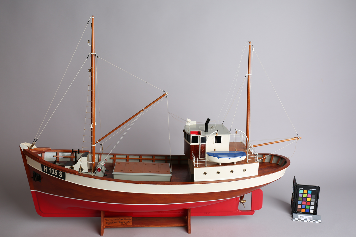 Modell av fiskeskøyte med to master. bygget i tre, med en livbåt. Står på stativ av tre. Detaljer som lanterner, skipsklokke og koøyer i messing.